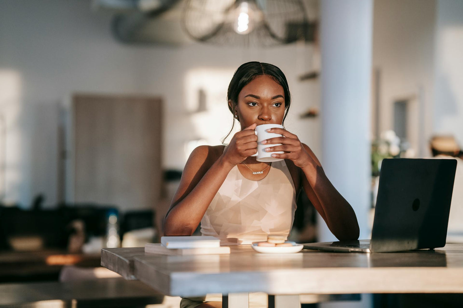 Woman drinking from a mug in a kitchen with a laptop on the table