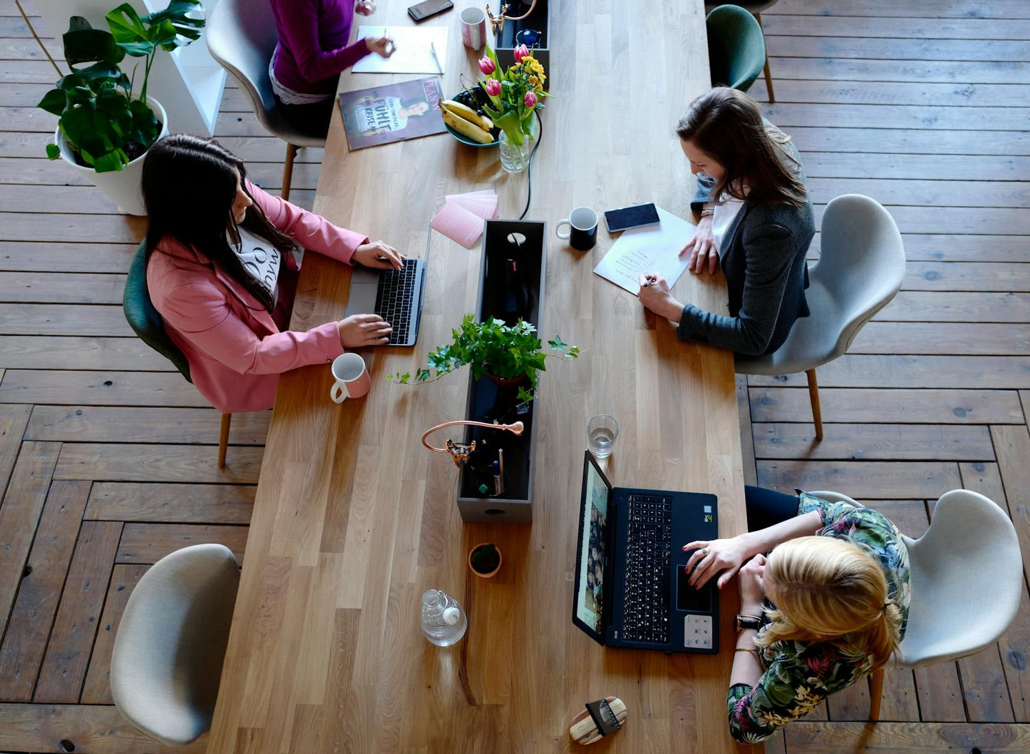 Three people working on laptops, drinking coffee at a wooden table in a modern office setting.