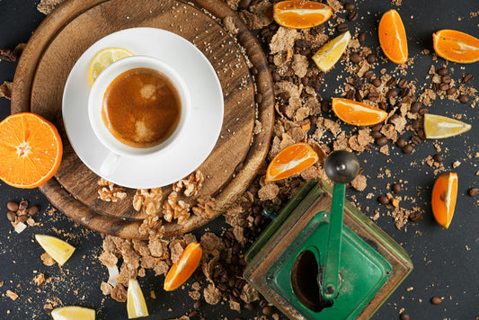 A cup of specialty coffee surrounded by oats, nuts, lemon wedges, orange slices, and a manual coffee grinder 