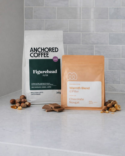 Anchored Figurehead Filter and Monogram Warmth Filter Blend coffee bags on a kitchen counter with hazelnuts, toffee and milk chocolate